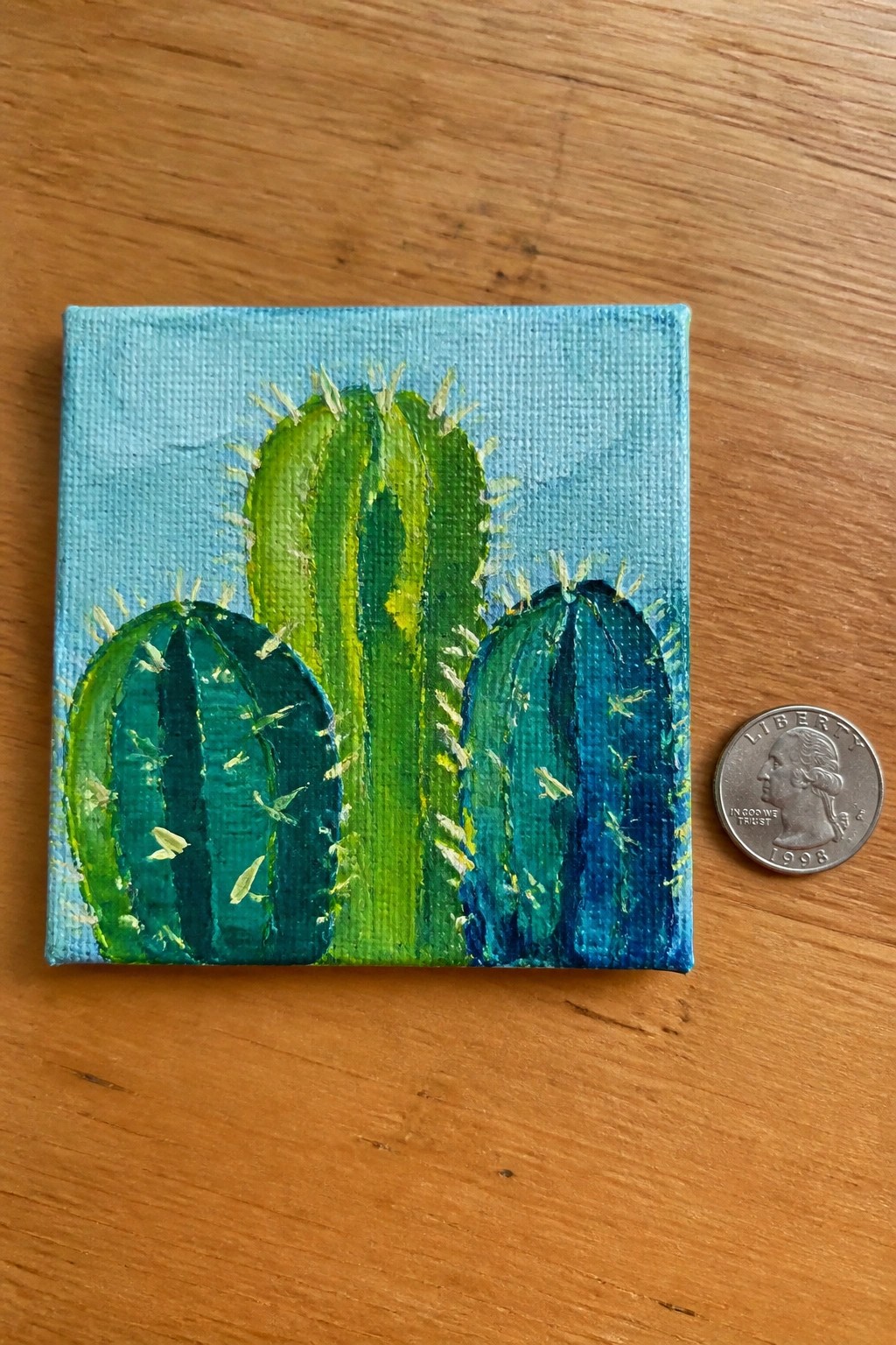 Small canvas painting of three blue-green cacti beside a 1998 US quarter on wood