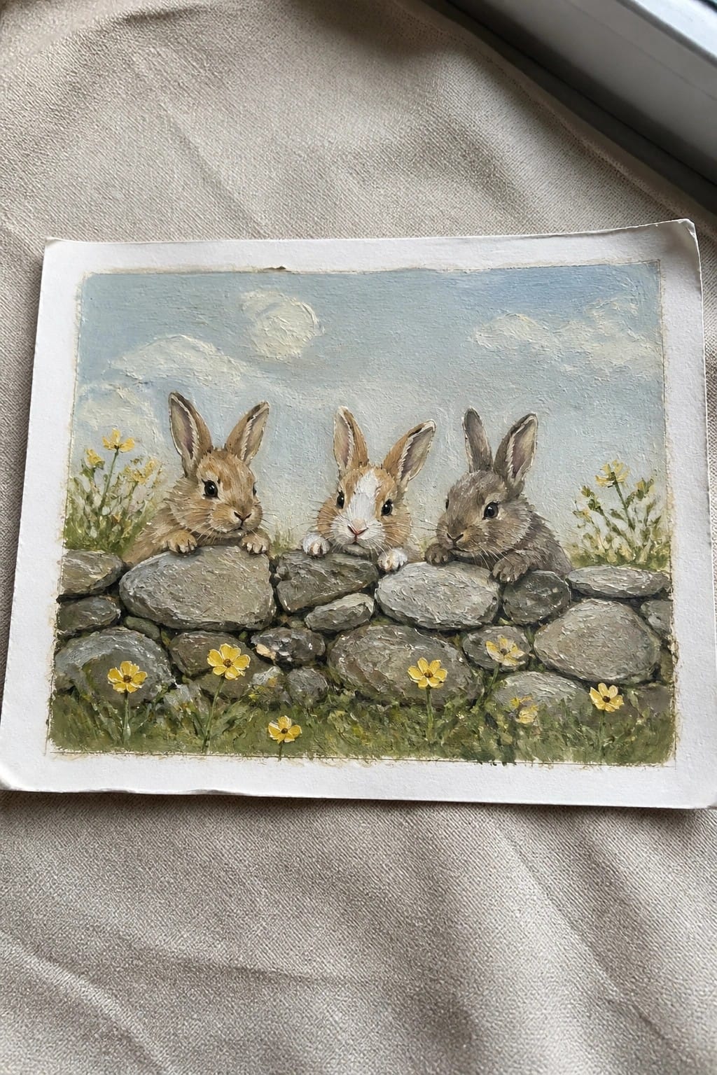 Three rabbits peeking over a stone wall amid yellow flowers under a blue sky.