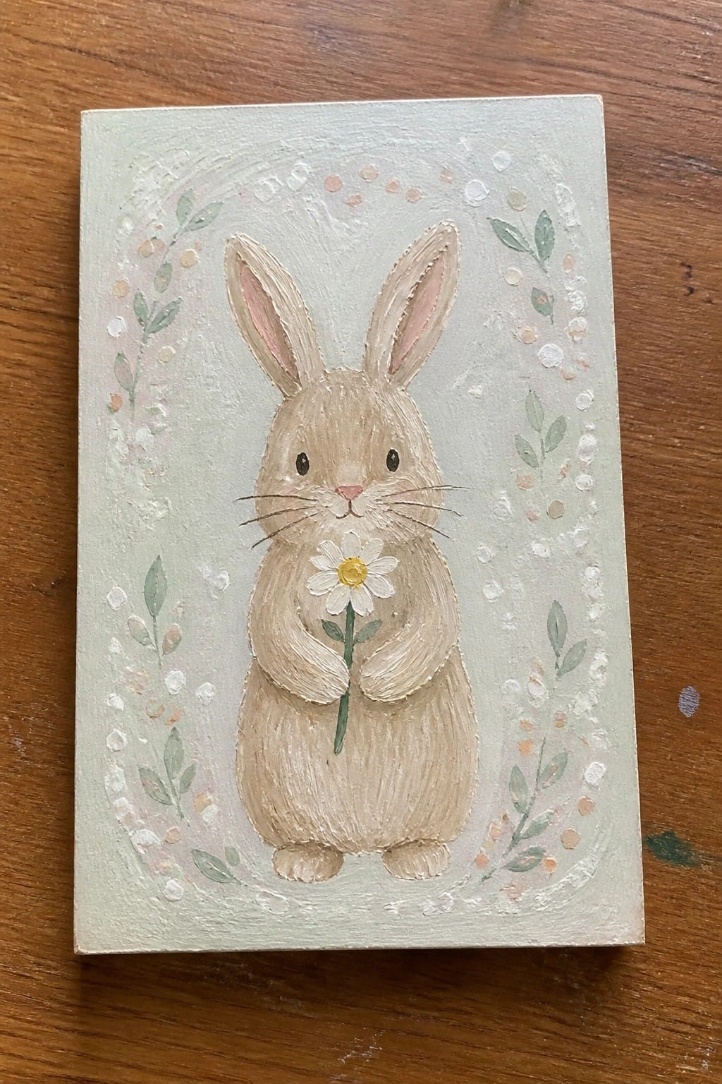Beige bunny with pink ears holding daisy on light green leafy background