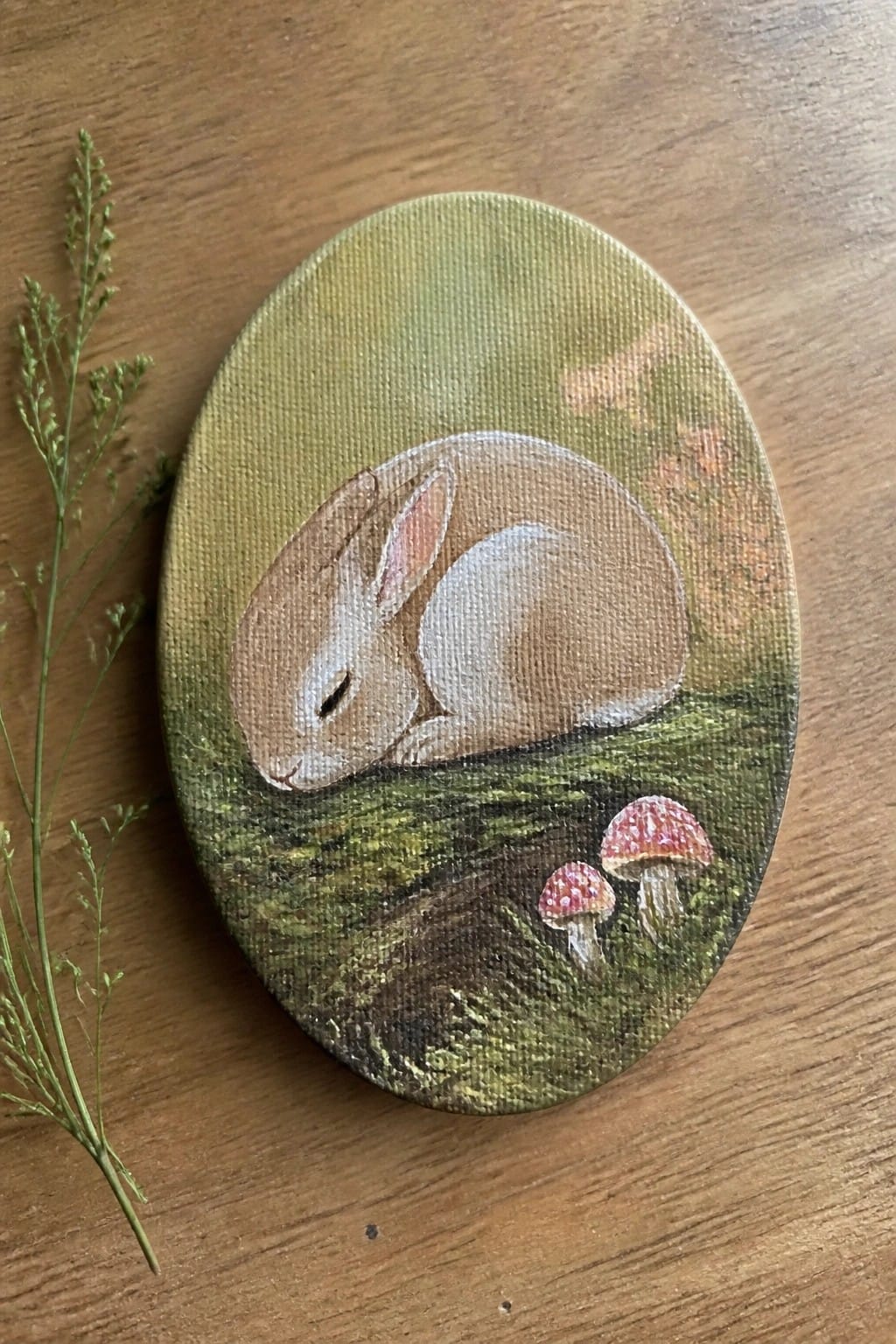 Oval canvas painting of sleeping brown rabbit beside red spotted mushrooms on wood.
