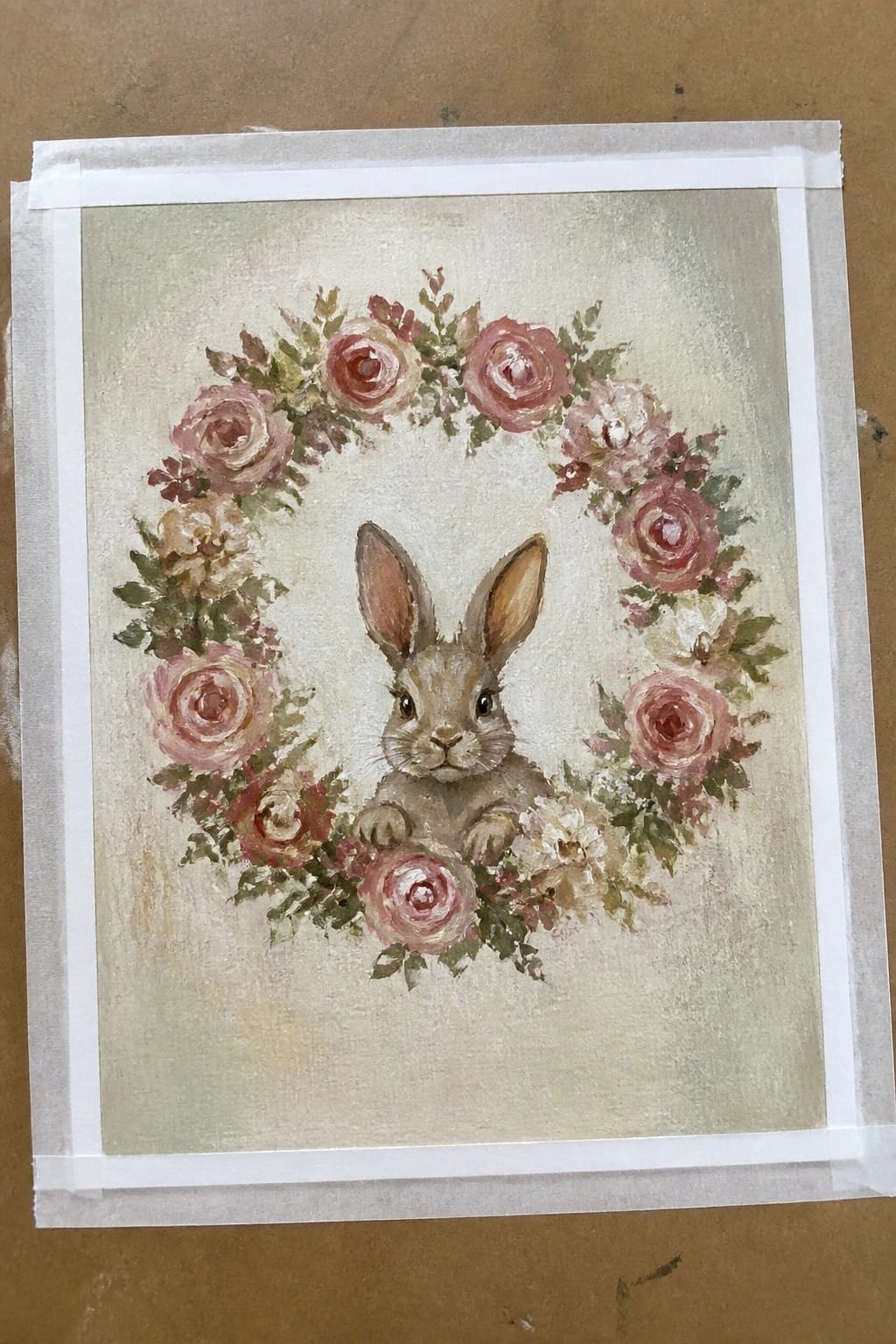 A painting of a small brown rabbit sitting inside a wreath of pink and white roses on a light background.