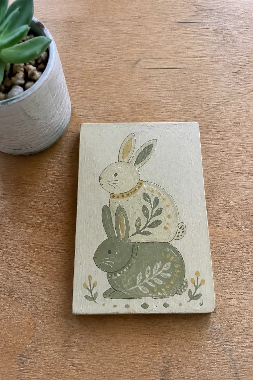 A rectangular wooden plaque shows two painted rabbits, one light beige above a green one, with leaves, dots, and small flowers around them.