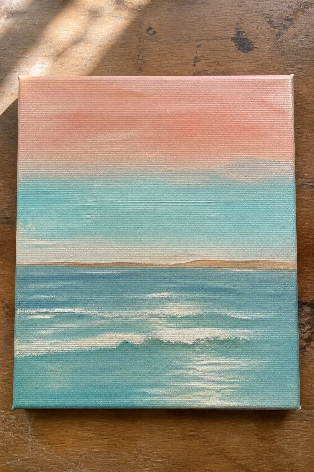 Small canvas seascape with turquoise waves, white foam, and peach-pink sky on wood.