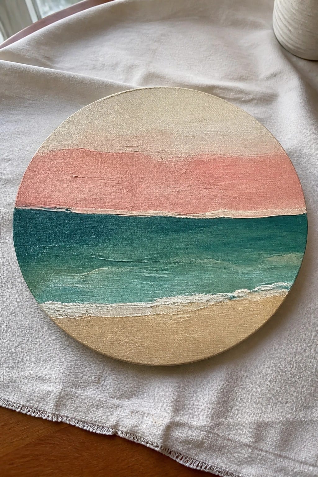 Round canvas painting of sunset over ocean with pink sky teal waves on white cloth