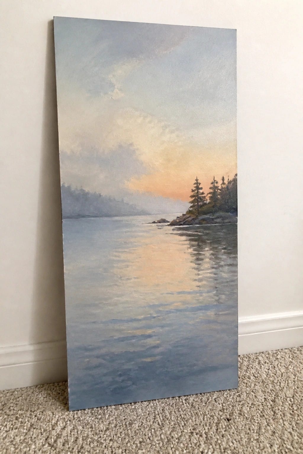 Landscape painting of calm water reflecting sunset hues over rocky pine-lined shore