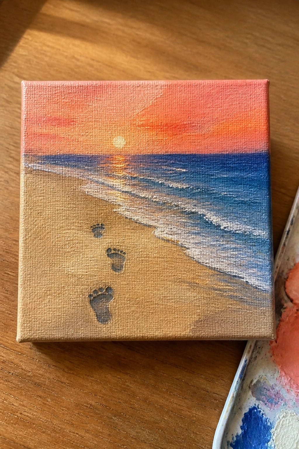 Square canvas painting of sunset beach with footprints in sand and ocean waves.