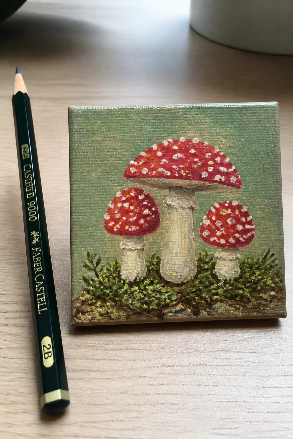 Miniature canvas painting of three red spotted mushrooms beside green 2B pencil on wood.