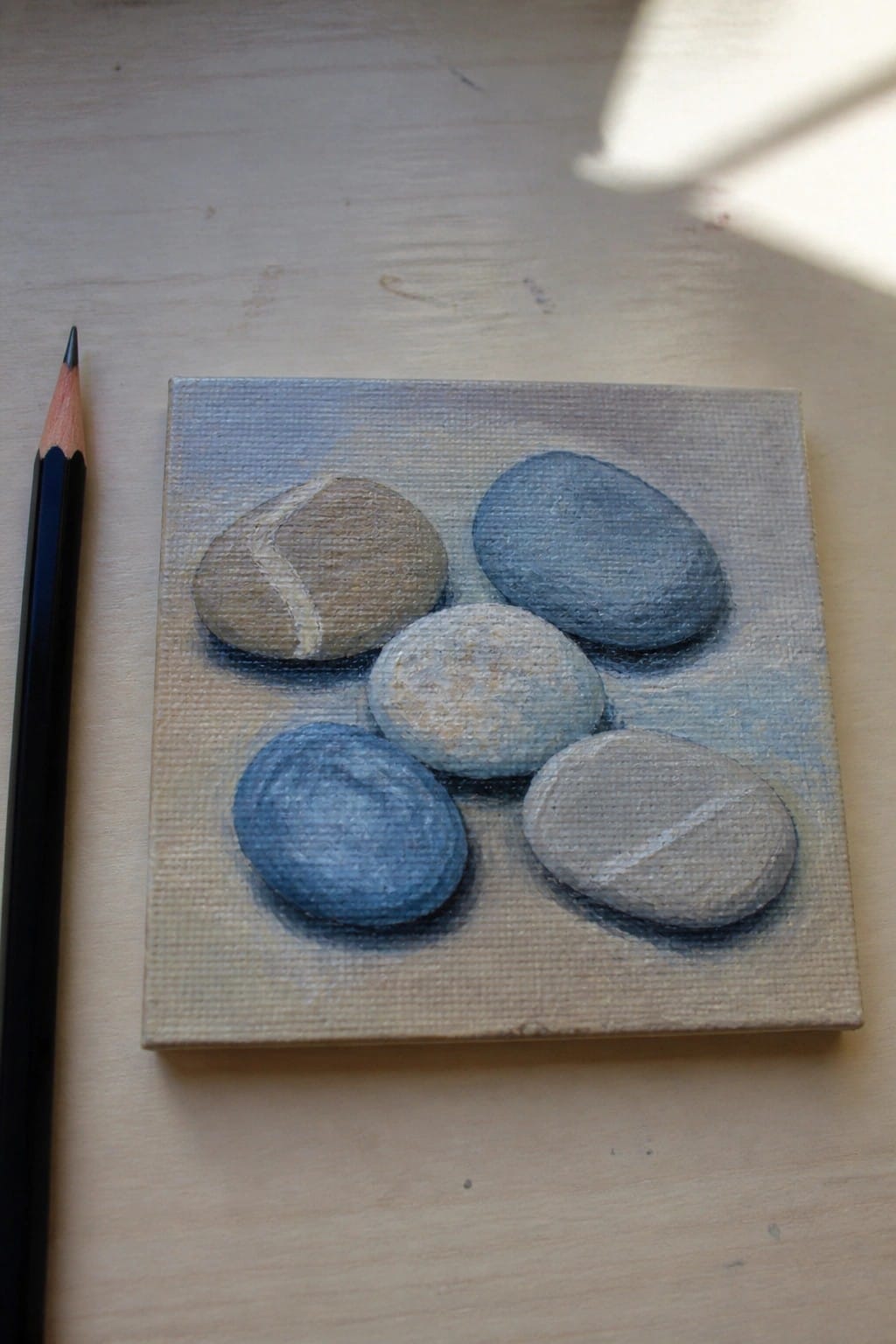 Small canvas painting of five blue-gray pebbles beside black pencil on wood