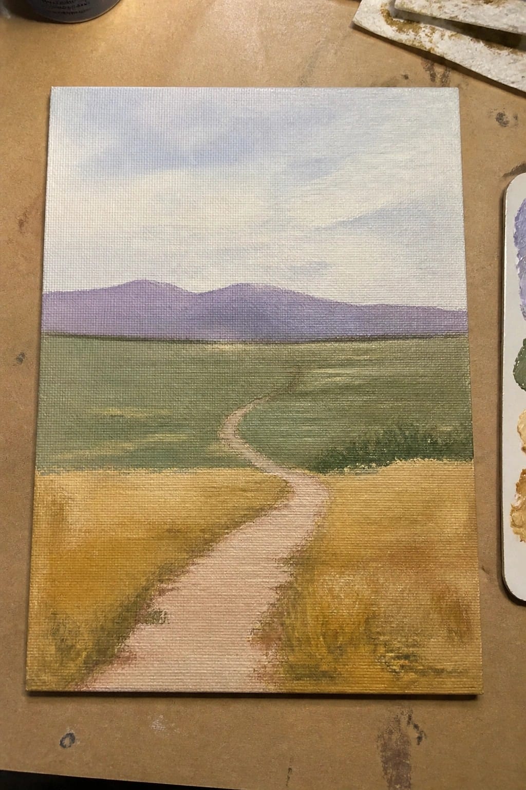 Winding path through green and yellow fields under purple mountains on canvas painting.