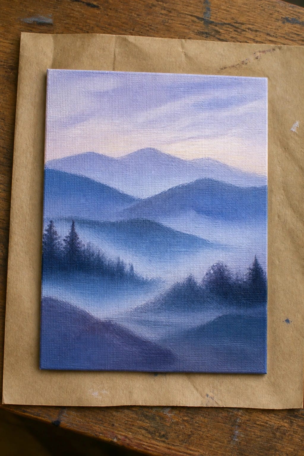 Mini canvas painting of misty blue mountains with trees on brown paper over wood.