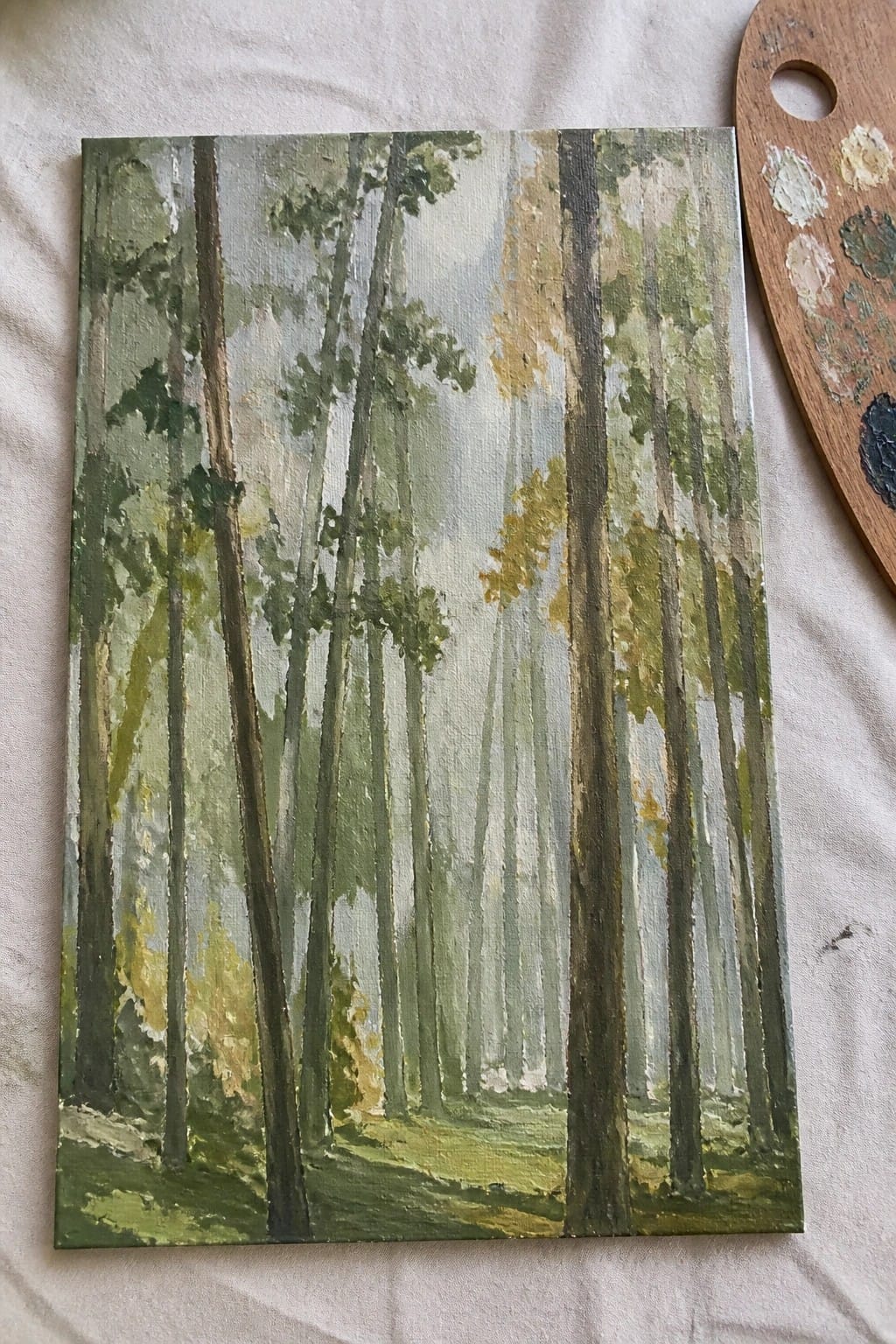 Oil painting of misty forest with tall green trees beside wooden palette.