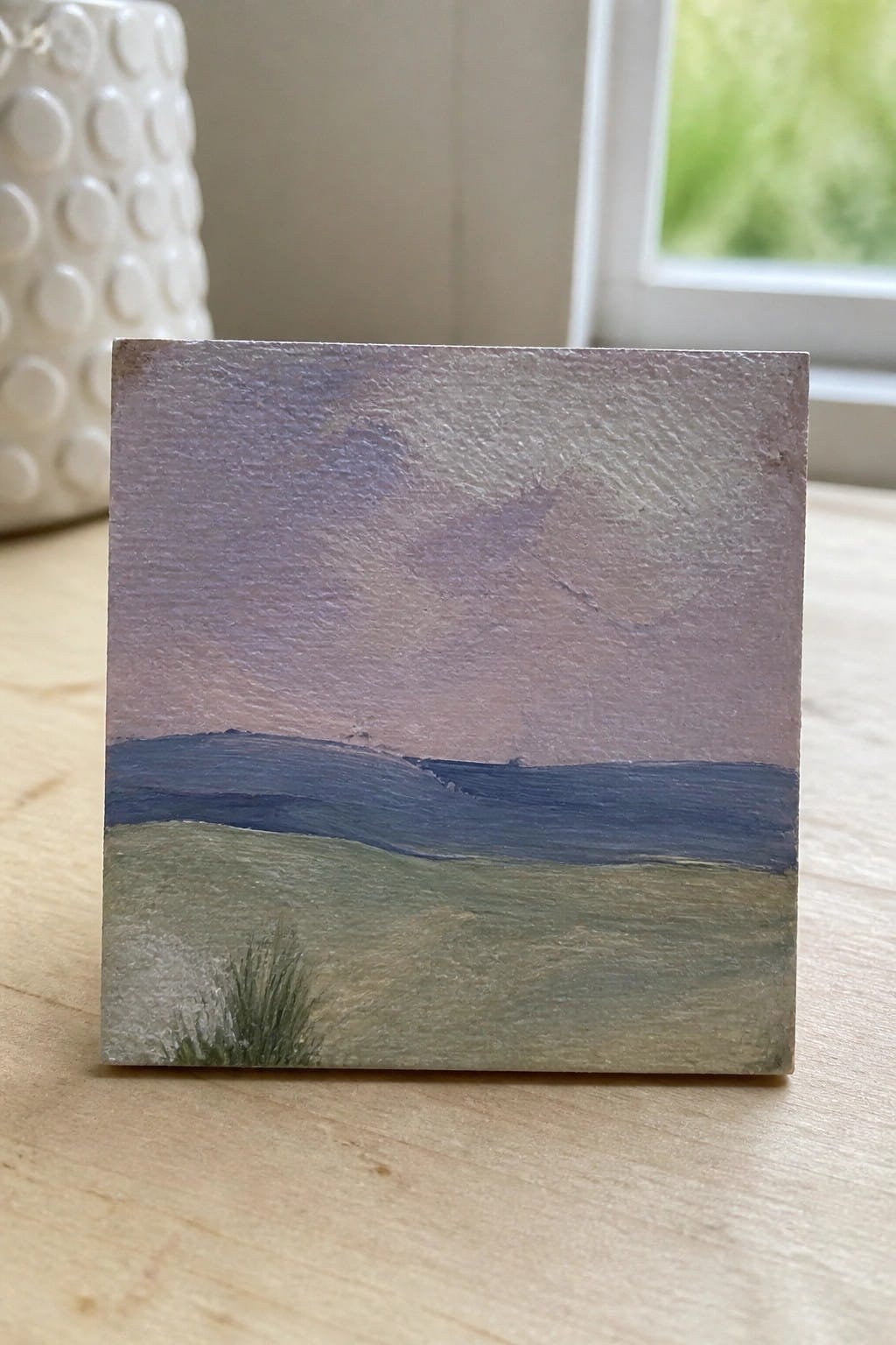 Small square painting of purple sky over blue hills and green fields on wood.