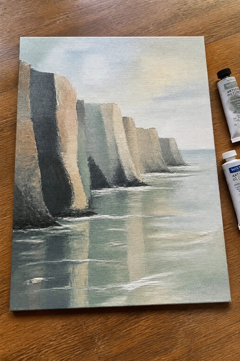 Oil painting of misty sea cliffs beside two tubes of paint on wood