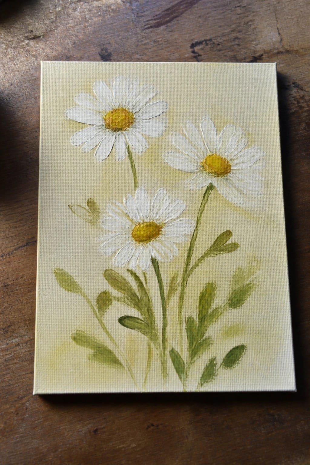 Three white daisies with yellow centers and green leaves on a canvas painting