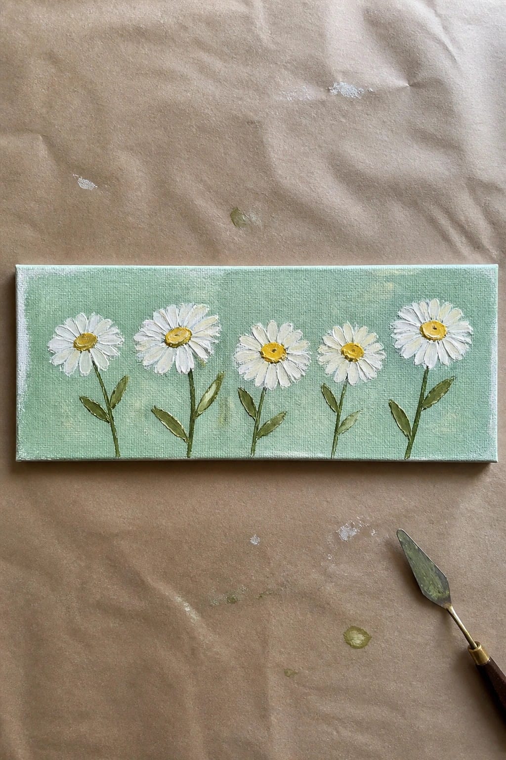 Five white daisies with yellow centers on light green canvas beside palette knife