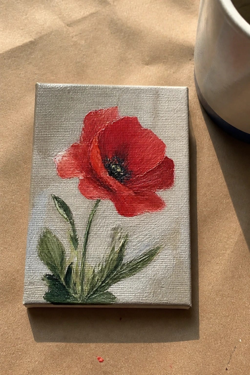 Small canvas with red poppy painting, green leaves, on brown surface