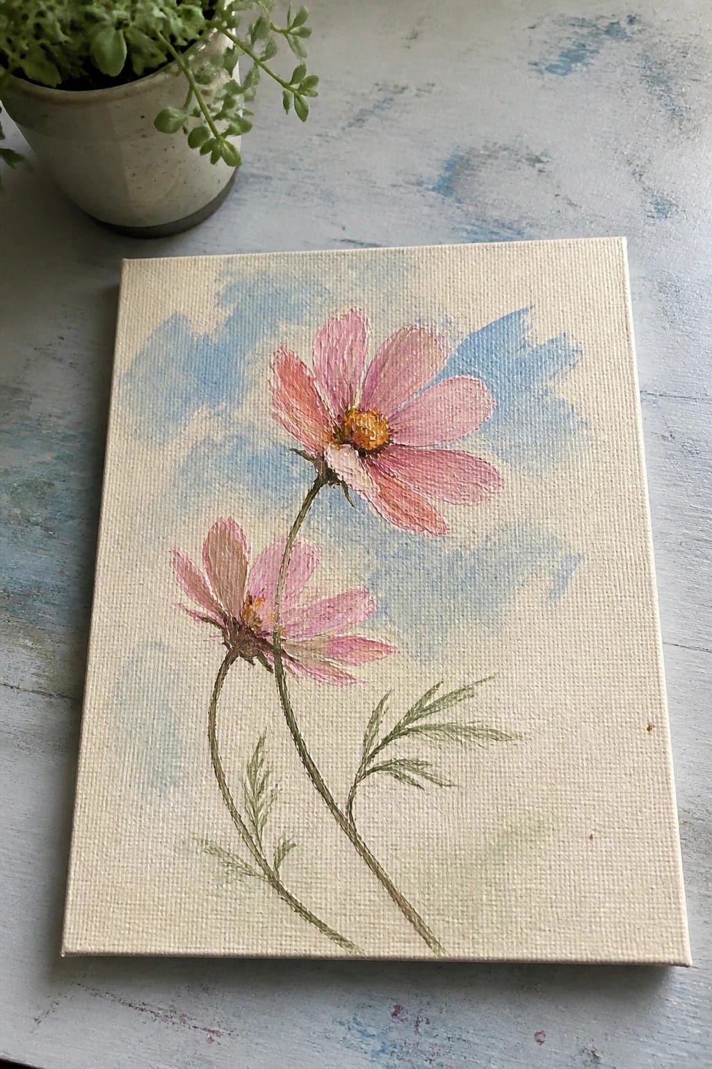 Pink cosmos flowers with green stems painted on canvas beside potted plant
