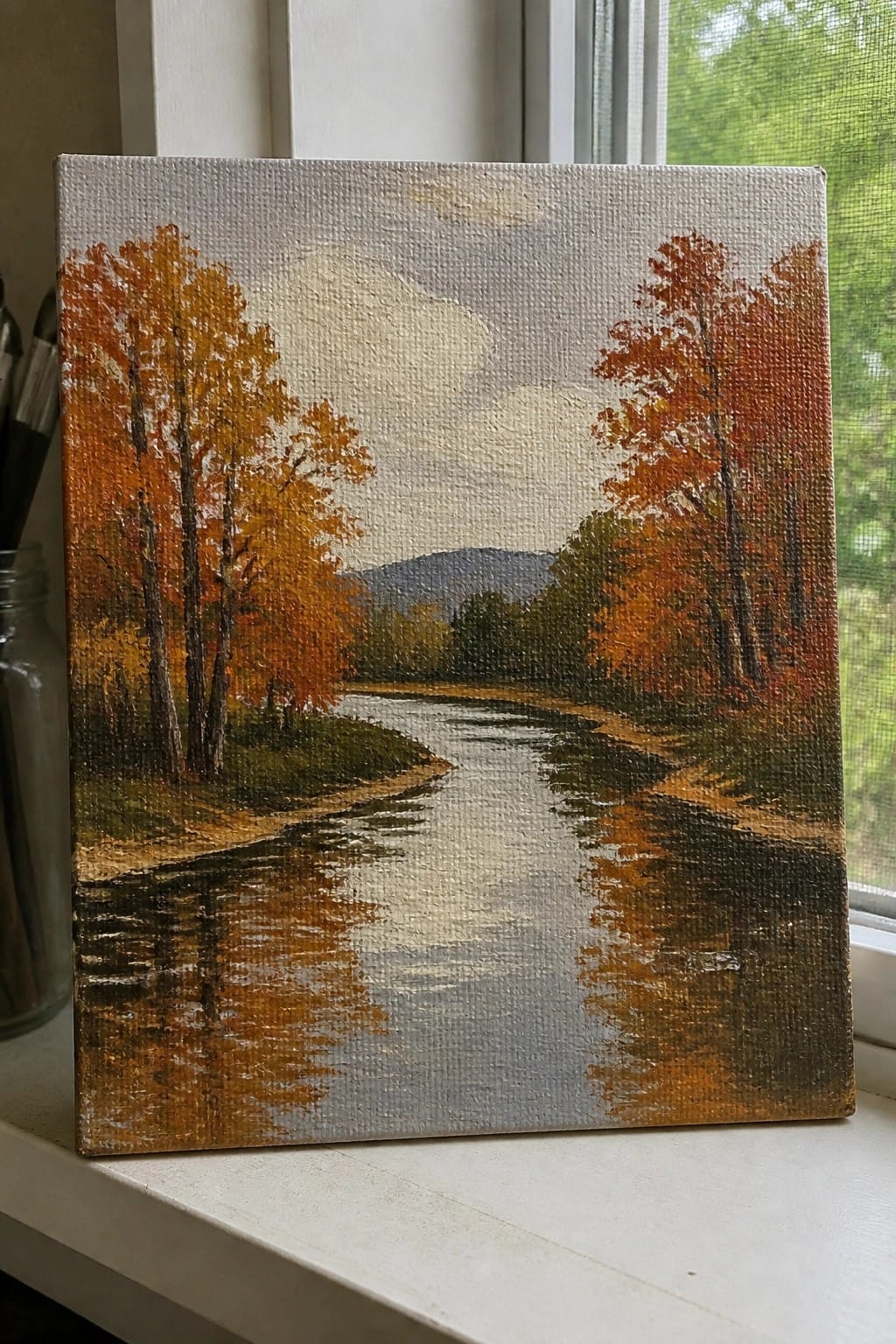 Painting of winding river reflecting orange autumn trees on windowsill