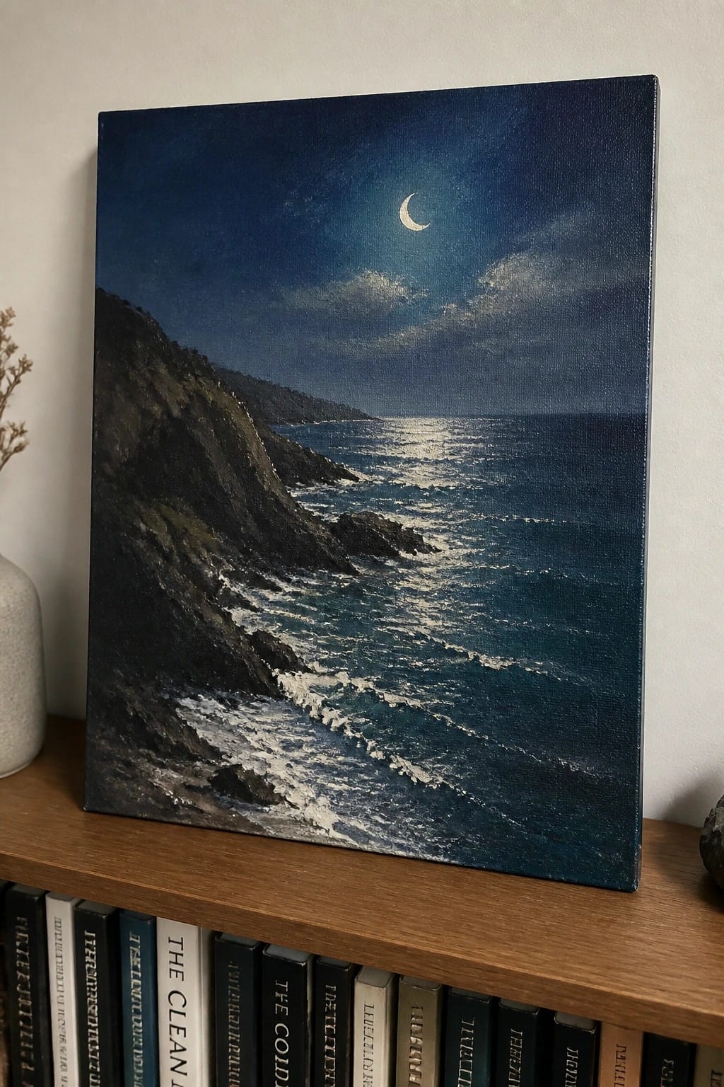 Dark blue seascape painting with crescent moon on wooden shelf above books