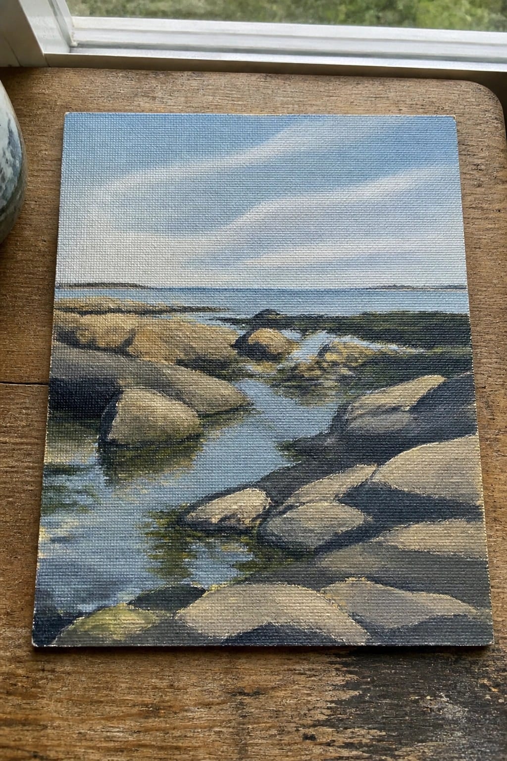 Small canvas painting of rocks in shallow blue water on wooden surface