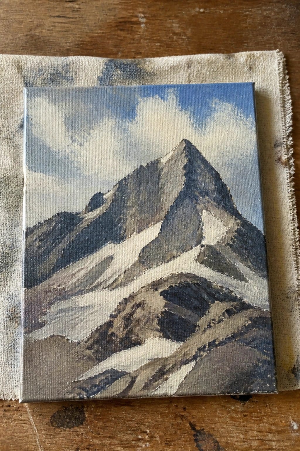 Small canvas painting of snow-capped mountains under blue sky with clouds on cloth.