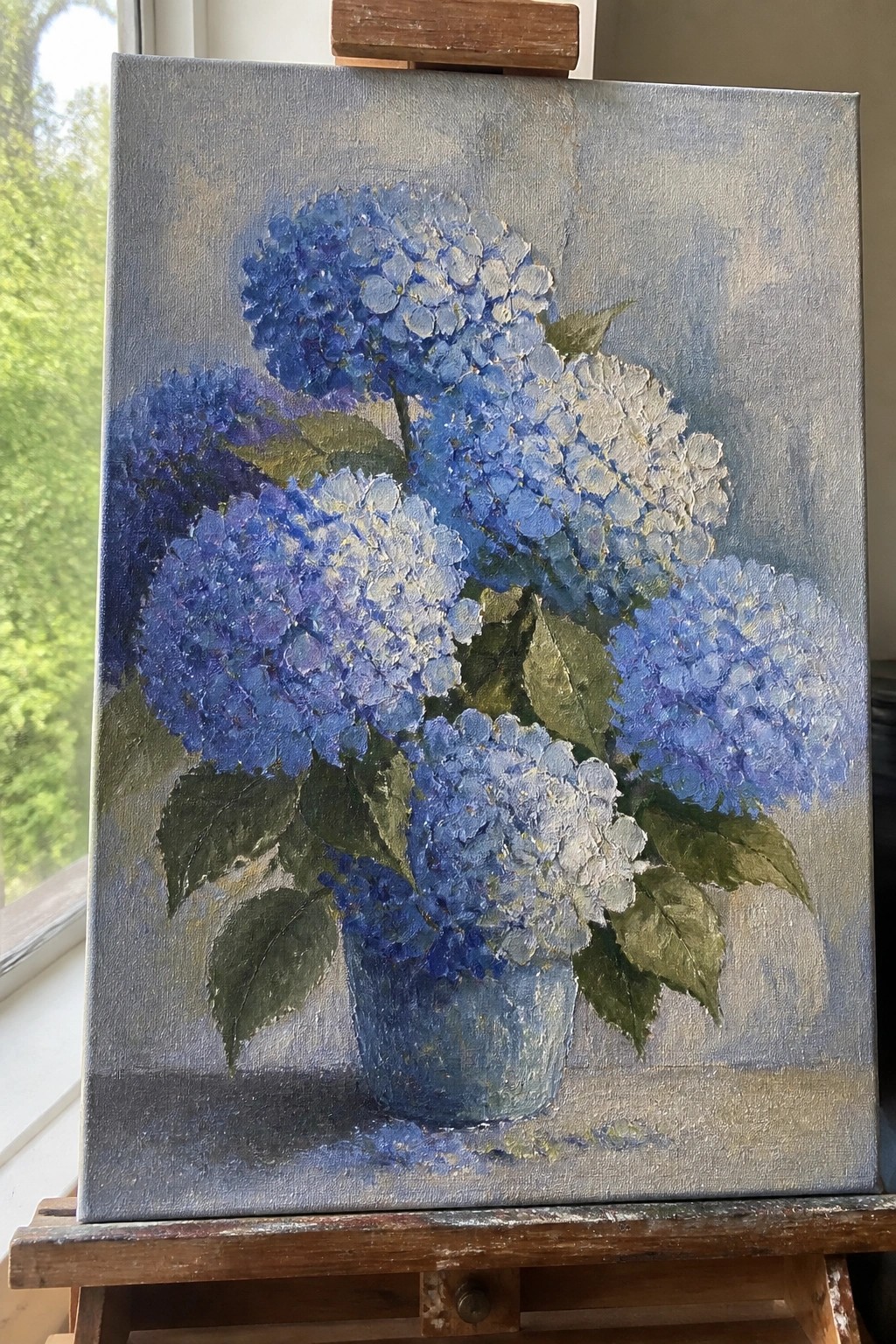 An oil painting of blue and white hydrangeas in a blue pot resting on a wooden easel.