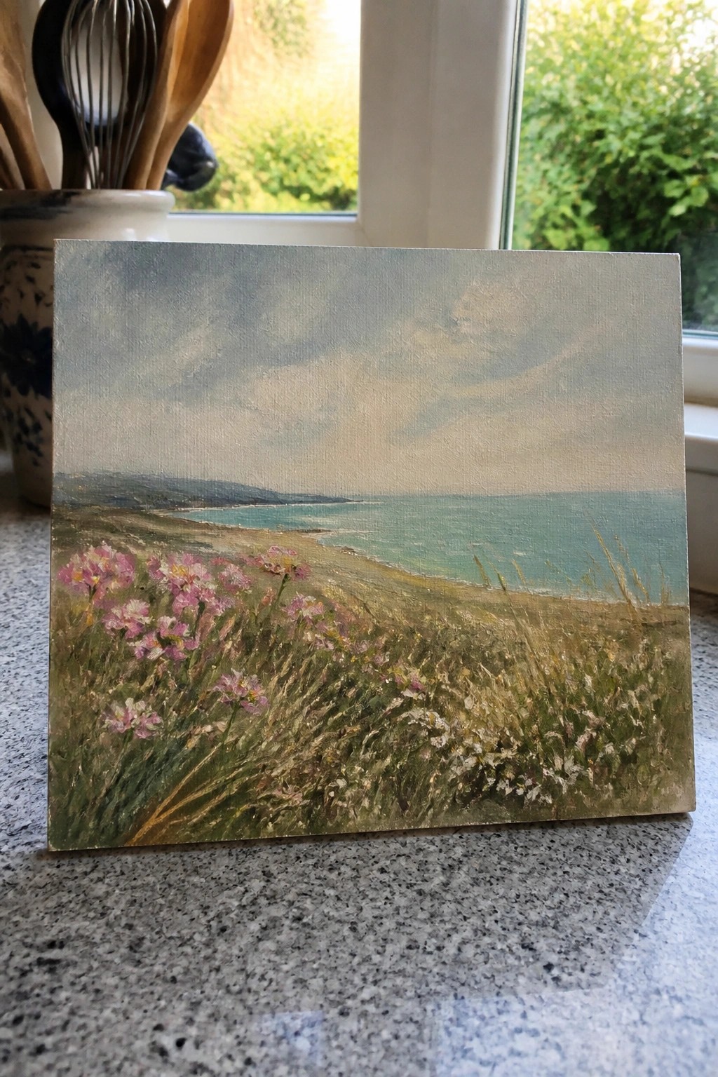 Small painting of pink flowers by turquoise sea on speckled kitchen countertop near window