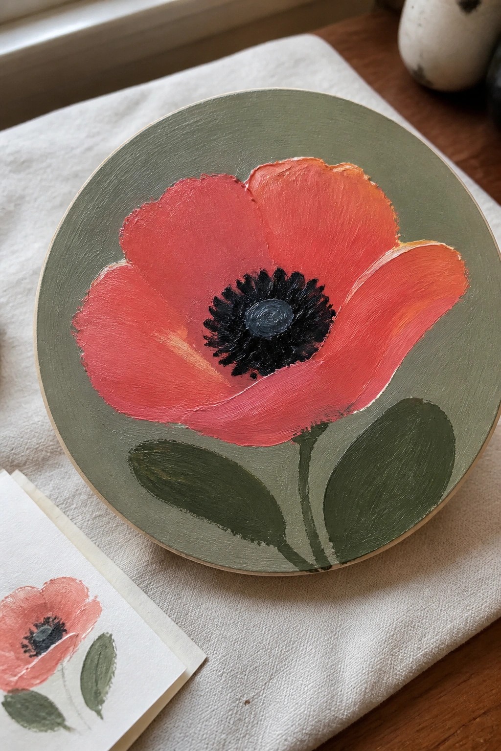 Red poppy with black center painted on round sage green wood beside sketch