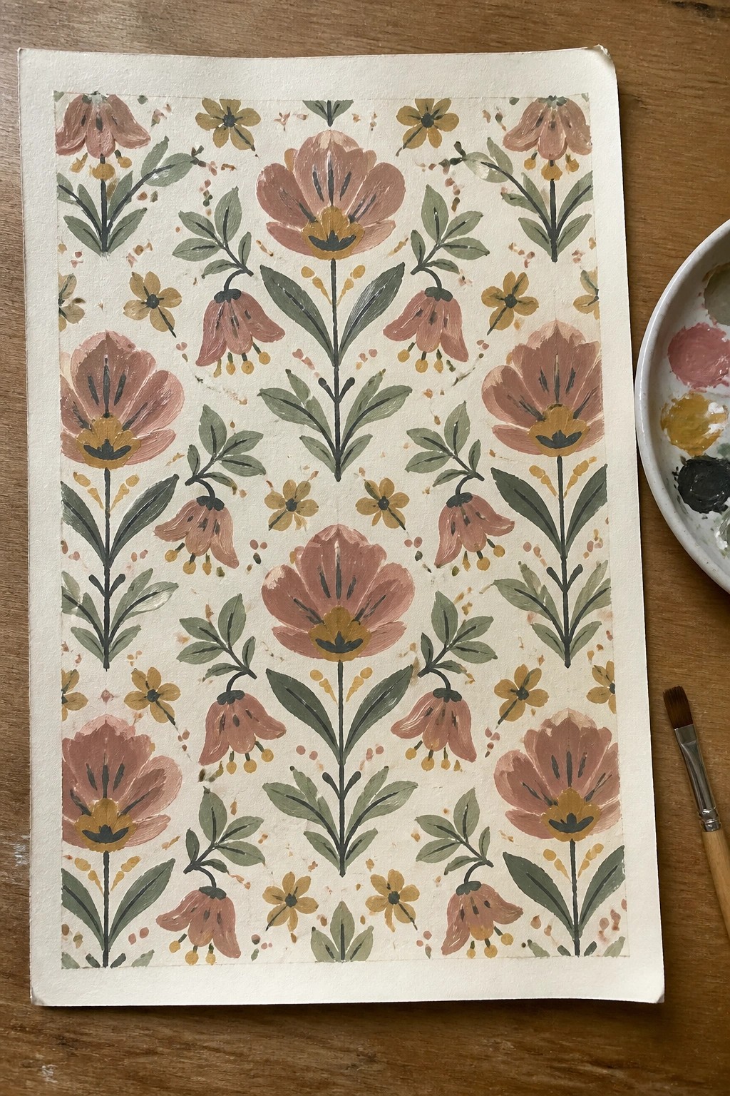 Watercolor floral pattern on paper with pink blooms, leaves, palette, and brush.