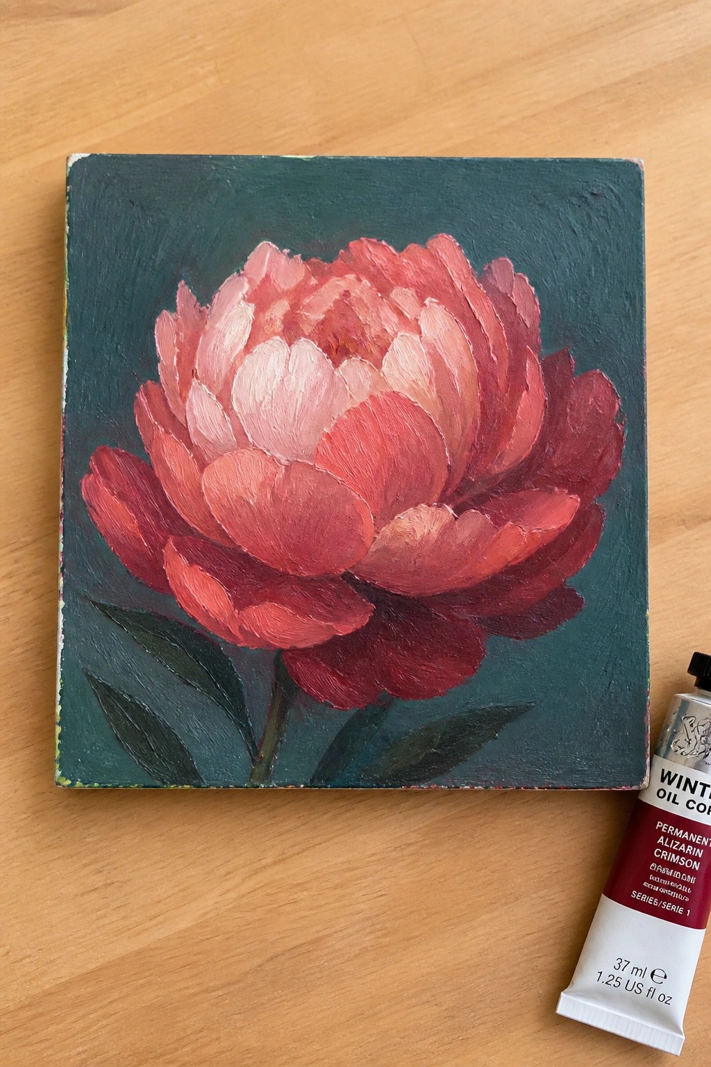 Oil painting of red peony on dark green canvas beside crimson paint tube