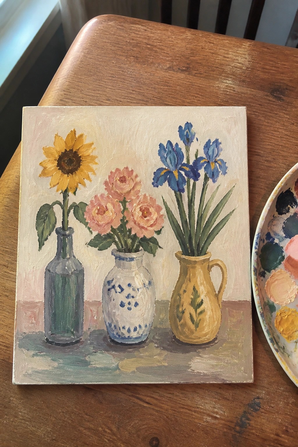 An oil painting of a sunflower in a glass bottle, pink roses in a white and blue vase, and blue irises in a yellow pitcher arranged on a wooden table next to a paint palette.