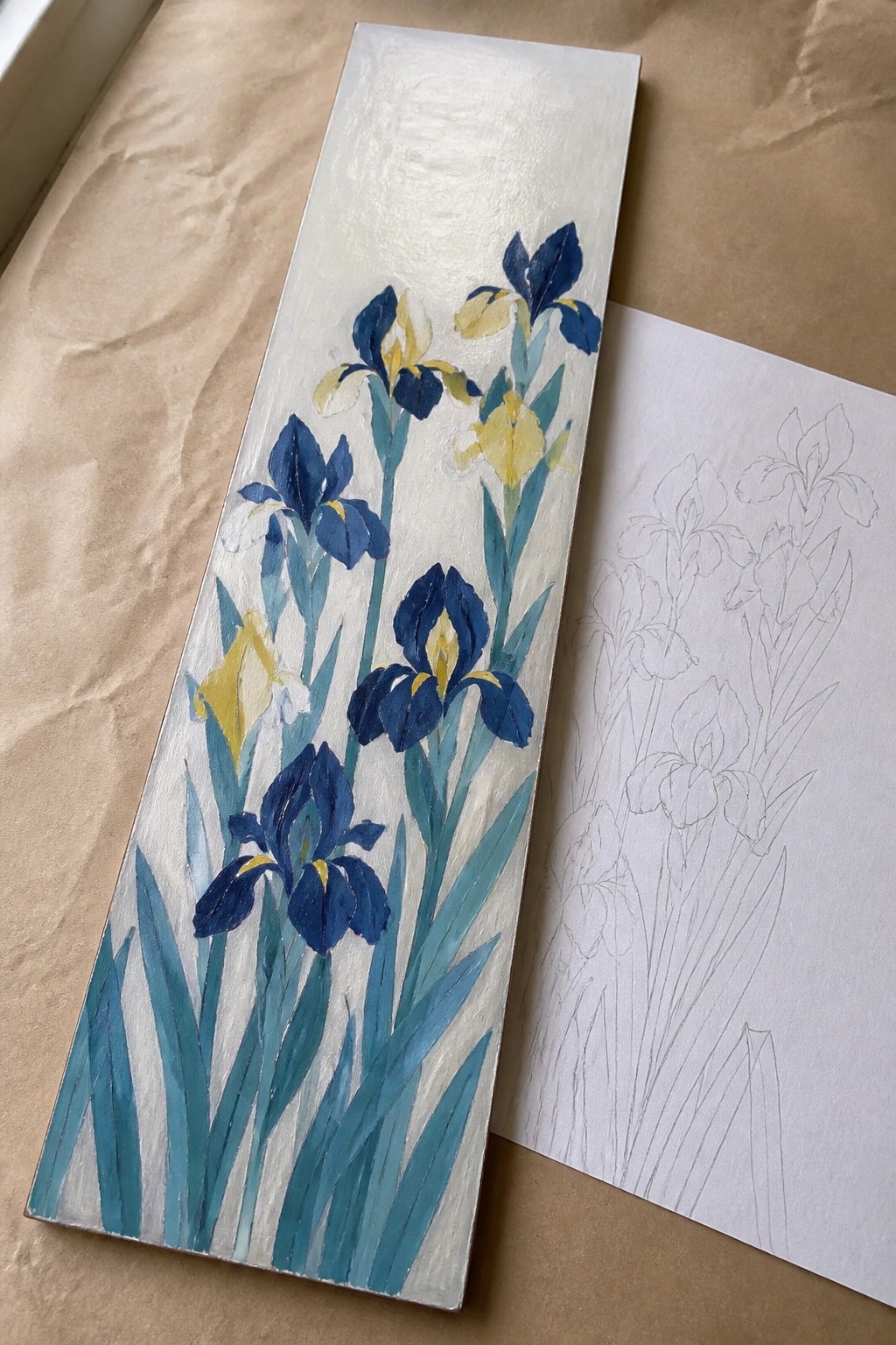 Narrow vertical oil painting of blue and yellow irises with green leaves on a white background next to a pencil sketch of irises on paper.