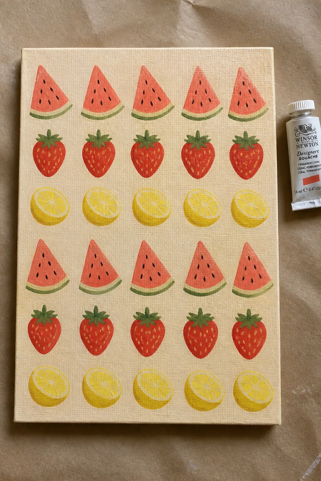 Canvas with rows of watermelon slices, strawberries, and lemon slices painted on a light background next to a tube of coral gouache.