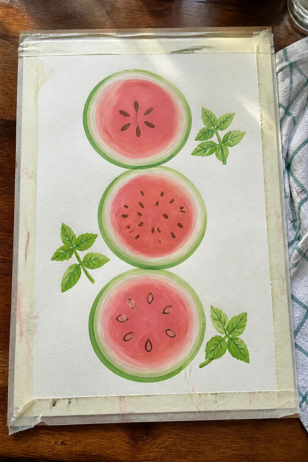 Three watercolor watermelon slices with green leaves on taped paper over wood