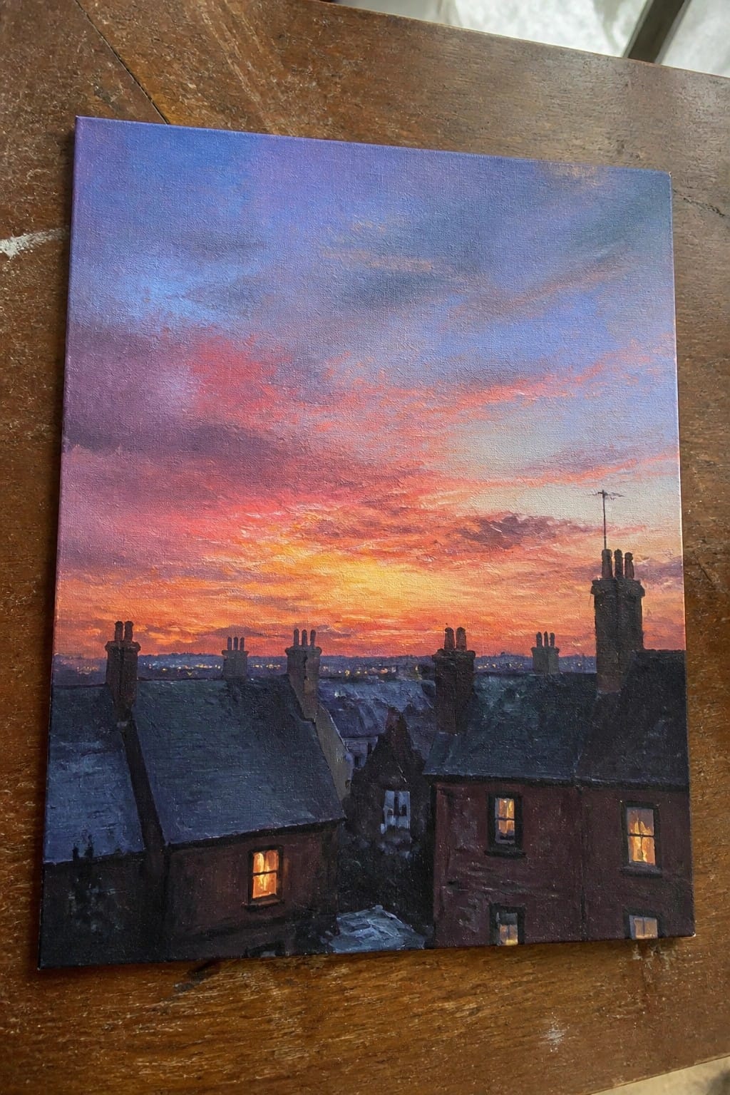 Oil painting of dark rooftops and chimneys silhouetted against a colorful sunset sky with a few lit windows.