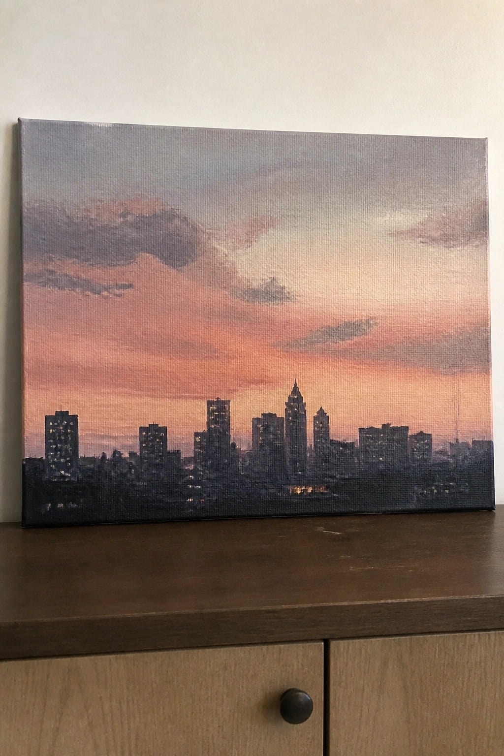 City skyline painting with glowing lights under pink-orange sunset sky on wooden dresser