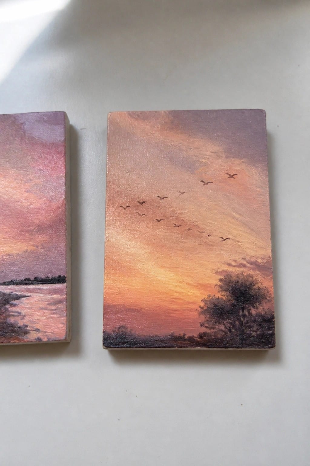 Two small sunset paintings, one over water and one with birds and a tree.