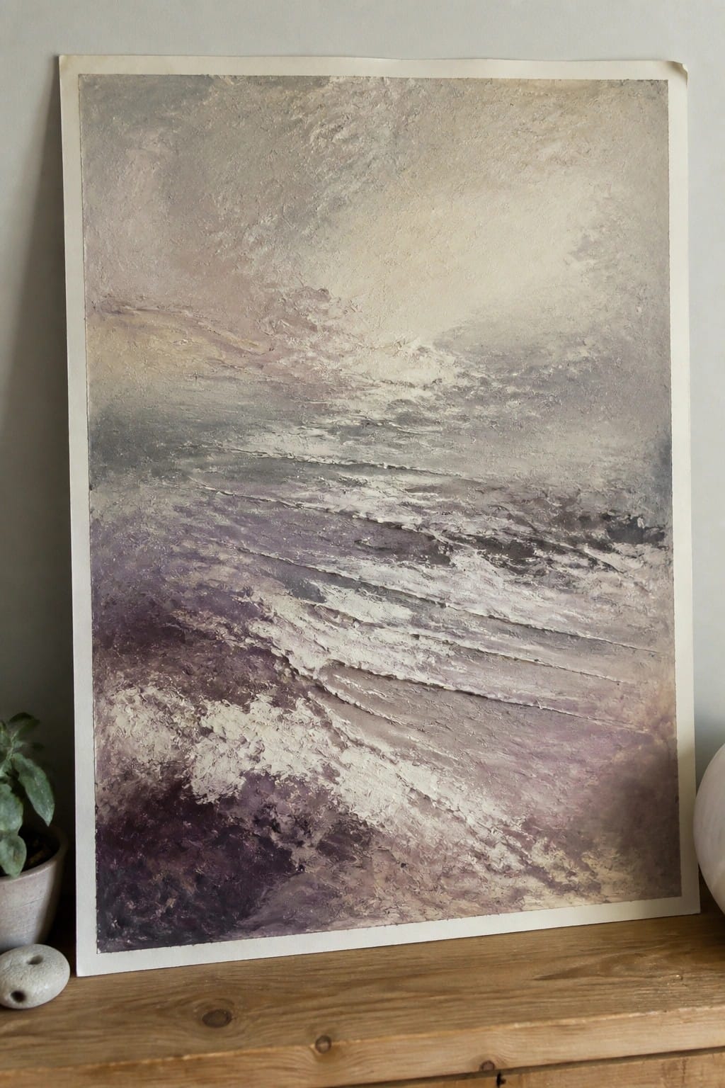Textured abstract painting in purple, gray, and beige tones on wooden surface with plant