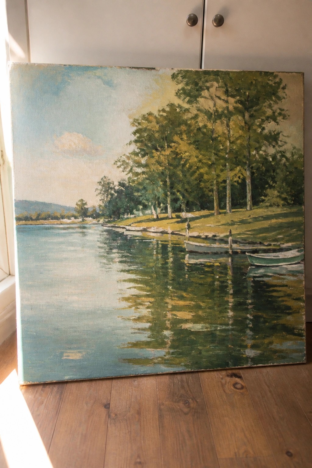 Large landscape painting of lake with boats and trees on wooden floor