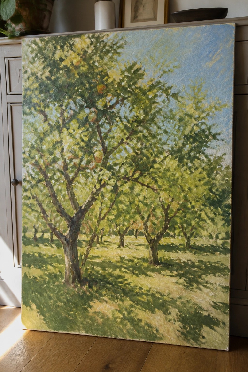 Impressionistic painting of sunlit apple orchard with green trees and blue sky indoors