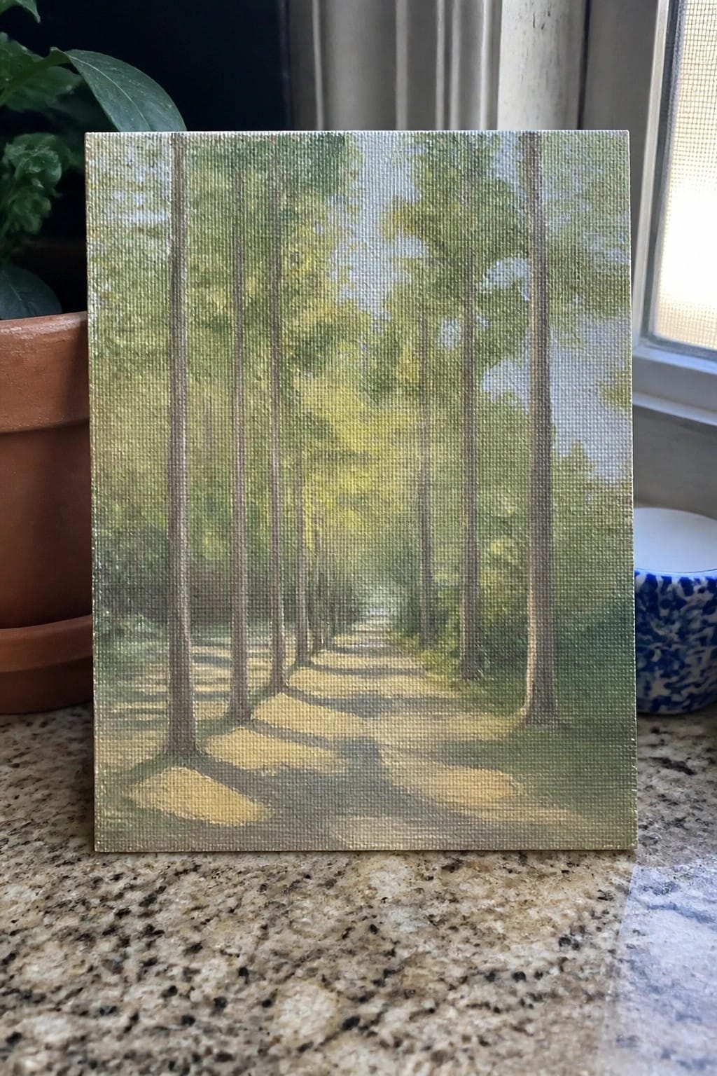 Small canvas painting of sunlit forest path with tall trees on granite countertop.