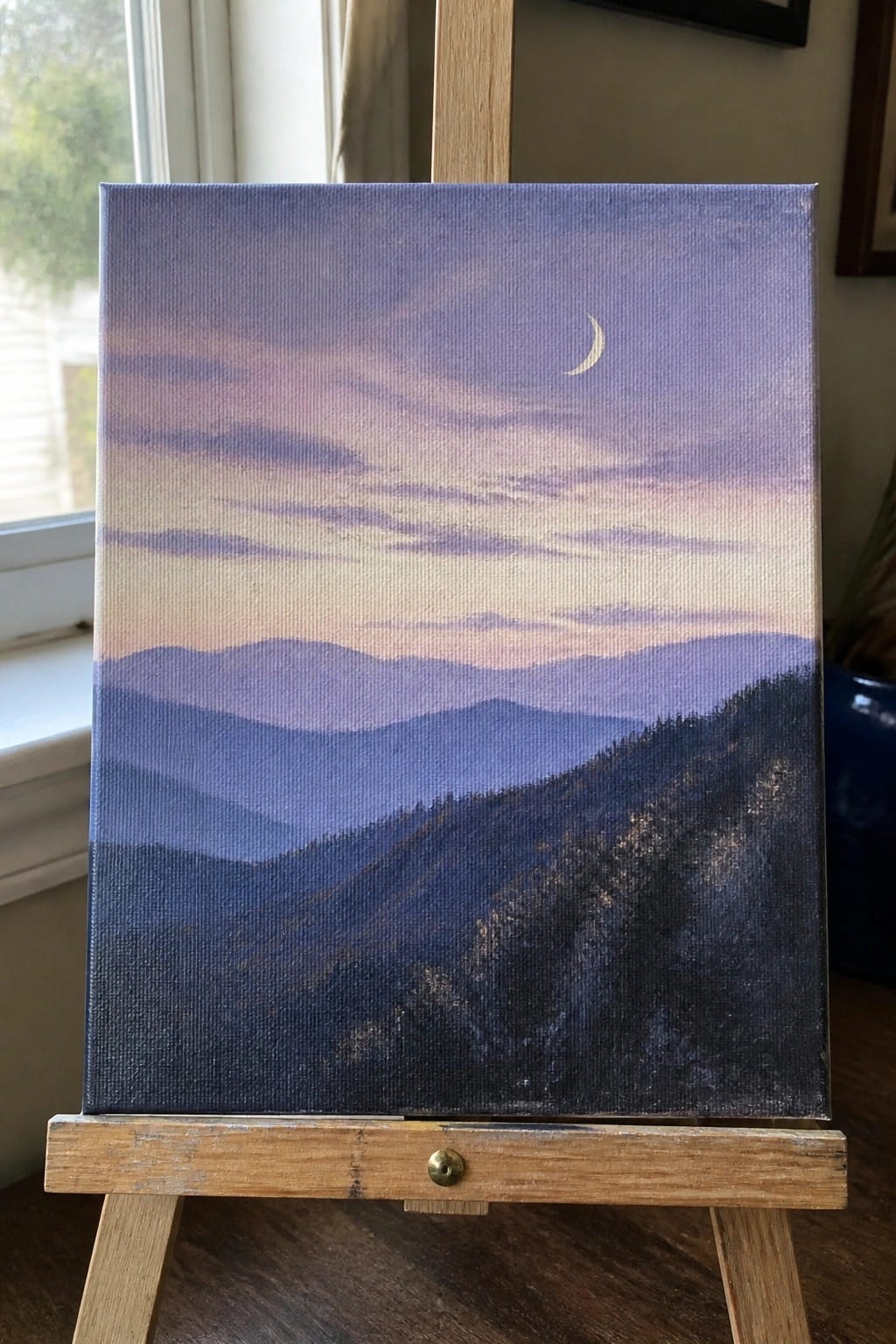 Canvas painting of layered blue mountains under purple sky with crescent moon on easel