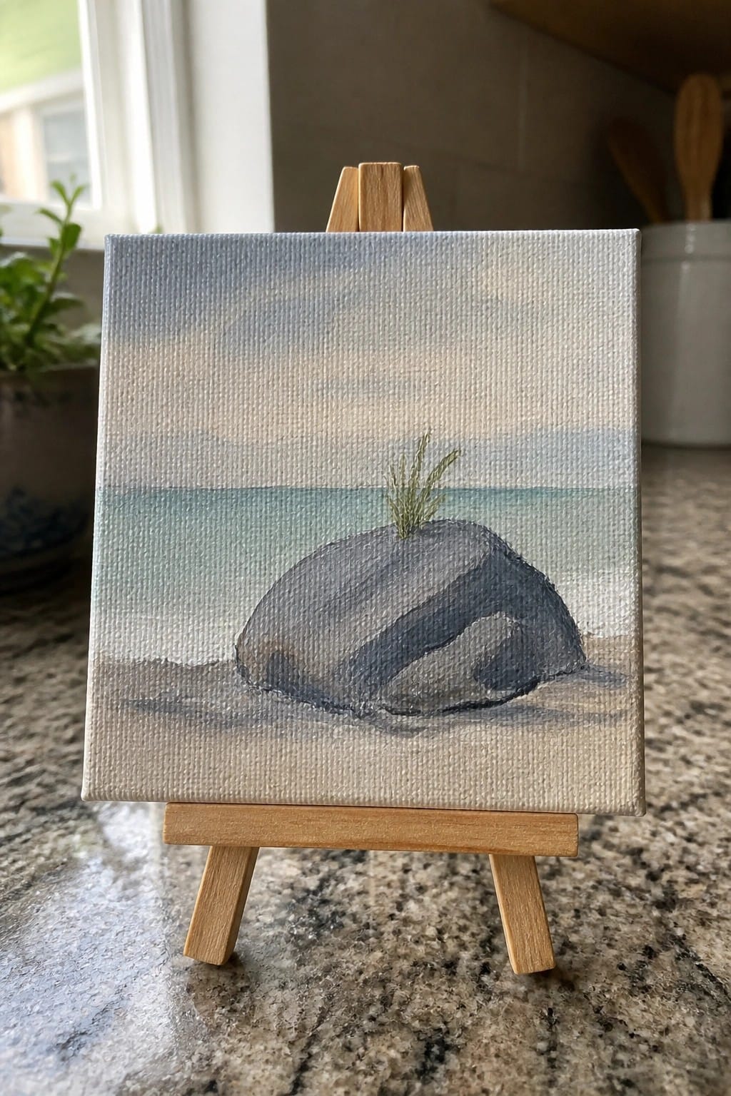 Miniature painting on wooden easel of a dark rock with grass near calm water
