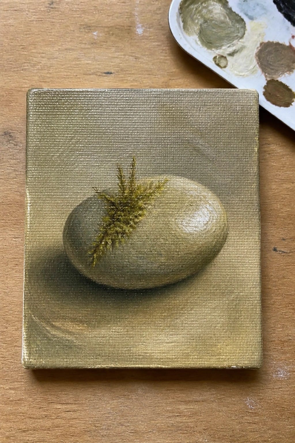 A small square oil painting shows a rounded greenish stone with a fine moss sprig resting on a textured earthy background next to a paint palette.