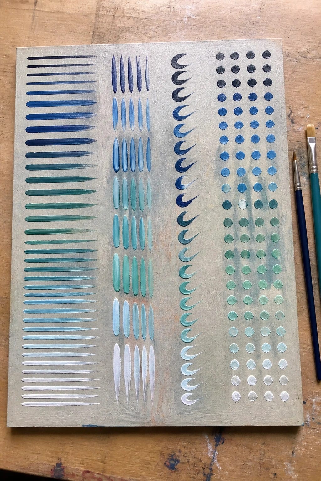 Canvas with blue gradient lines, ovals, crescents, and dots beside paintbrushes on wood
