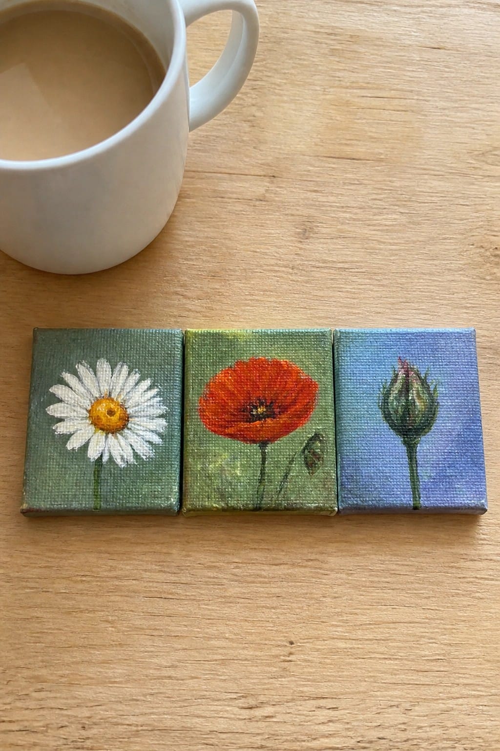 Three miniature flower paintings—a daisy, poppy, and bud—beside white coffee mug on wood