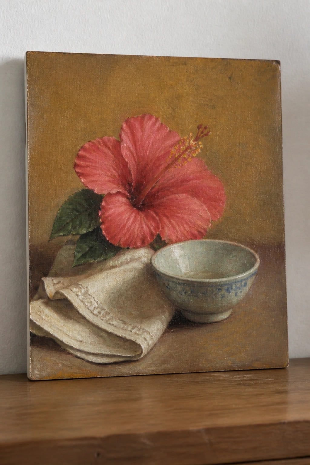 Still-life painting of red hibiscus flower beside patterned bowl on folded cloth.