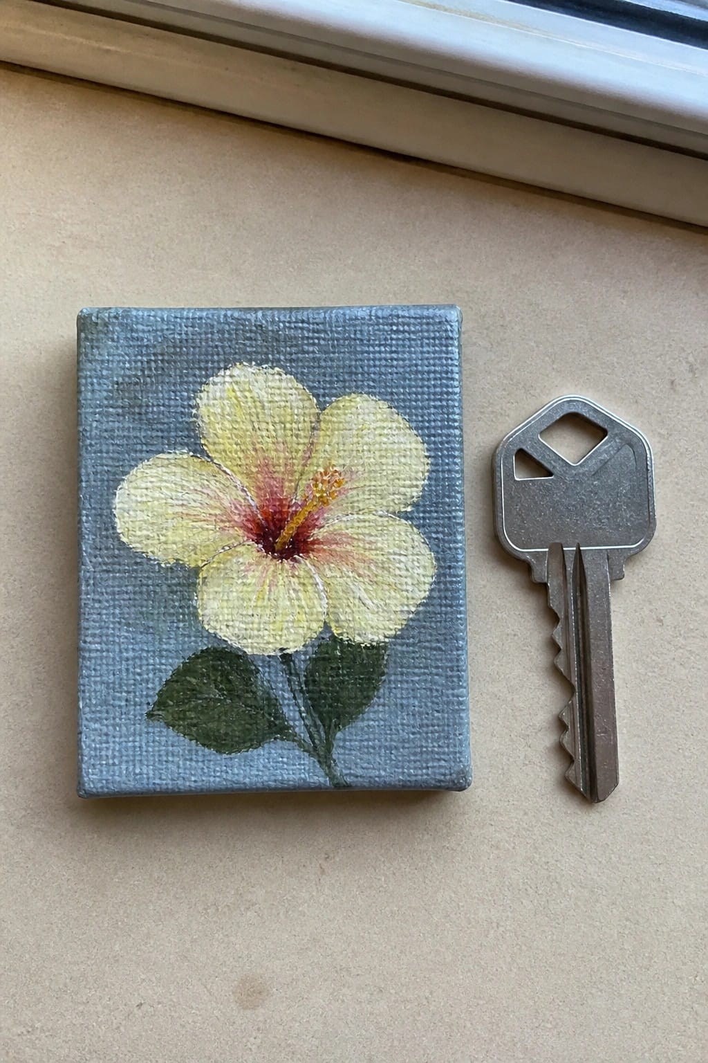 Small yellow hibiscus painting on blue canvas beside silver key