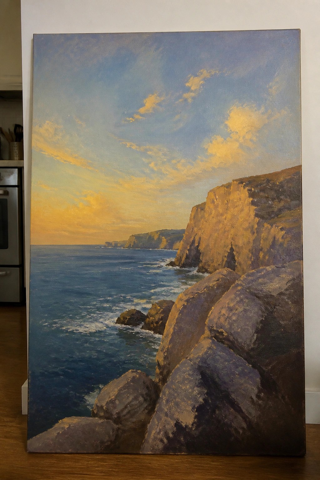 Large oil painting of rocky cliffs by blue ocean under golden sunset sky.