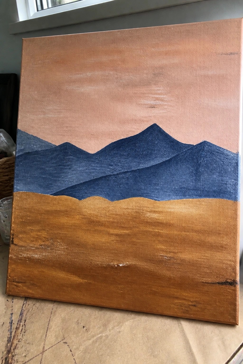 Canvas painting of layered blue mountains under peach sky with brown foreground